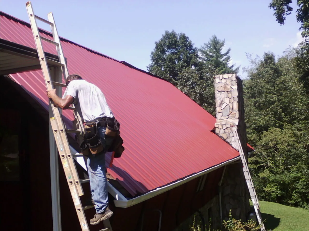 Roofer on a ladder beside a red metal roof and stone chimney — Soffit & Fascia Repair by Roofing Brothers in Whiteman Air Force Base